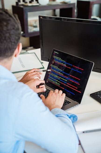 The image depicts a person working at a desk with a laptop displaying lines of colorful programming code. The individual, seen from behind, is using a stylus or pen to interact with the device, which 