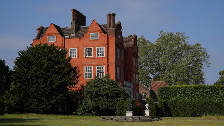 A large red brick Victorian-style house features multiple stories, with distinctive gabled rooflines and several brick chimneys extending from the roof. In front of the house, a well-maintained garden